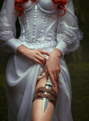 Fantasy red-haired dangerous woman medieval assassin queen with dagger in sheath on leg leather belt. White vintage old style dress. Girl princess warrior. Green summer nature trees. Hand hold knife
