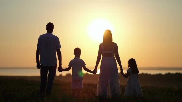 Happy family looks at sunset standing on huge meadow. Mother and father silhouettes raise hands with kids to heaven looking at sunset against blue sky