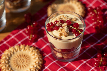 Dessert im Glas mit Johannisbeeren, Vanillecreme und Keks auf einer roten Tischdecke im Sonnenlicht