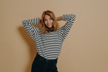 Half length shot of pretty satisfied young woman keeps hands behind head smiles gently poses indoor