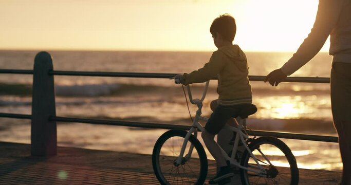 Learning to ride a bike with a father teaching his son to cycle at the beach by the sea. Young boy child on his bicycle with his dad, having fun and bonding together as a family at sunset in summer