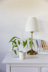a branch of eucalyptus in a glass vase, a box of books, a massage roller for the face, a cup of coffee on a white chest of drawers against the wall. Ready layout. Vertical frame. Space for text
