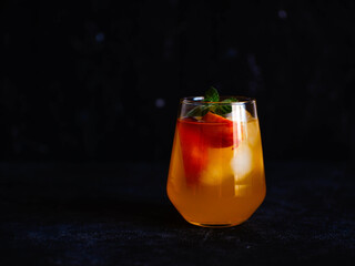 A glass of peach cocktail with ice, mint leaves on a dark background.