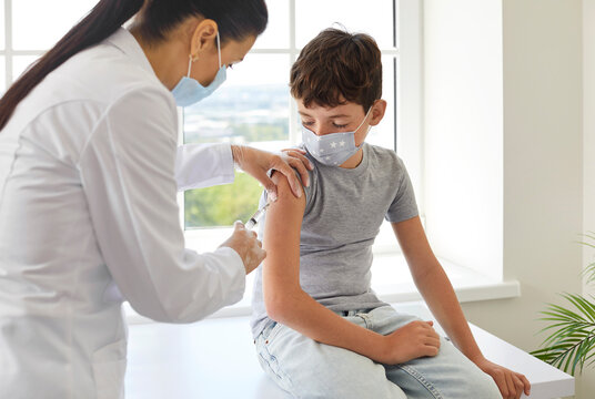 Nurse Or Doctor Gives Shoulder Injection To Little Boy To Protect Him From Dangerous Disease. School Child In Medical Facemask Gets Flu Or Covid 19 Vaccine At Clinic During Mass Vaccination Campaign