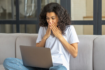 Fototapeta na wymiar Pleasantly shocked young African American woman looking at screen of laptop, gets good news sitting on the couch at home is excited from happiness, success concept
