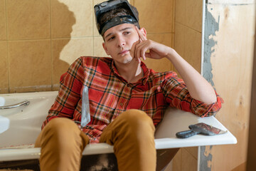 a young man, a plasterer or painter, sits in a bathtub with a spatula in his hands and looks at the...