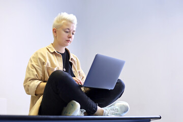 Girl with short blonde hair sitting on office table cross-legged with laptop in her hands. Tomboy...
