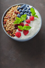 Granola with yogurt and berries on a dark brown background. Healthy breakfast with yogurt, baked granola, blueberries and raspberries. Top view. Copy space