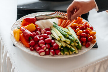 A hand reaches for a bowl of vegetables. On a plate are tomatoes, cucumbers, carrots, pepper, radishes. Useful and healthy food.
