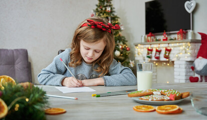 Child girl writes Letter for Santa in Christmas room. Christmas cookies and glass of milk on table. Funny Christmas time and holidays.