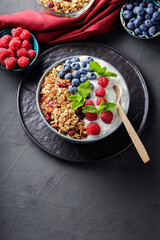 Breakfast cereal granola with berries and greek yogurt on a dark background. Bowl with yogurt, granola and fresh berries on a black plate. Healthy breakfast with wooden spoon