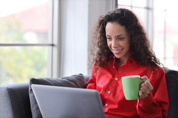 Smart and active latin woman sitting on sofa and using internet on computer with smart mobile phone while drinking coffee in living room. Lifestyle with technology