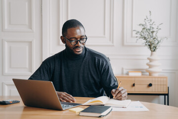 Focused young african american businessman writing down notes in notebook while working in office
