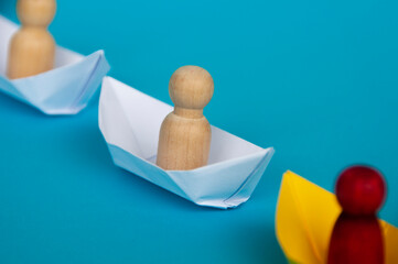 Wooden doll people figure on white paper ship origami following the red doll on yellow ship. Copy...