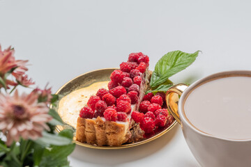 Top view of delicious piece of cake with fresh raspberries on golden plate. White background. Cup of delicious coffee and blurry chrysanthemums in the foreground. Sweet concept.  Place for your text.
