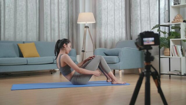 Full View Of Young Asian Trainer Female In Sports Clothes Speaking To Camera And Doing Crisscross Crunch Workout While Recording Teaching Exercise At Home

