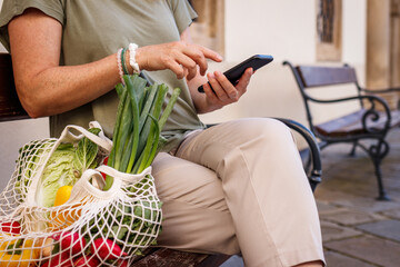Woman with reusable bag sitting on bench and using smart phone in city