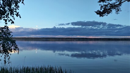 There are ripples in the water of the lake. Evening is coming. The moon has risen over the forest on the far shore and clouds have accumulated. Pine and birch branches lean over the water and reeds