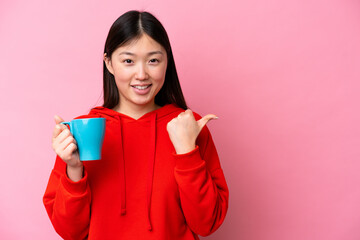 Young Chinese woman holding cup of coffee isolated on pink background pointing to the side to present a product