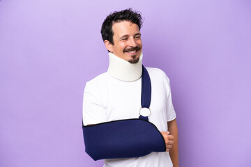 Young caucasian man wearing a sling and neck brace isolated on purple background looking to the side and smiling