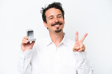 Young man holding a engagement ring isolated on white background smiling and showing victory sign