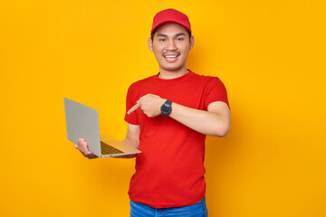 Happy young Asian man in red cap t-shirt uniform, employee work as dealer courier, standing with pointing fingers at laptop pc computer on yellow background. Professional Delivery service concept