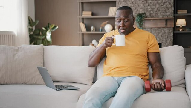 African sportsman american senior drink hot coffee burn tongue sit on sofa make domestic exercises stretch muscles raise dumbbell gymnastic workout program feel vitality energy sporty leisure at home