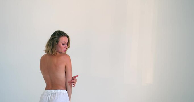 the girl turned to the camera with her bare back, stroking her shoulder and arm with her hand and looking into the camera. blond girl in white pants half naked. medium shot