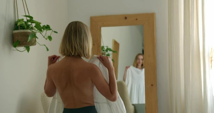 a blonde girl with a bare back looks into a wooden mirror and tries on a white dress. on the wall is a green flower in a vase. medium shot