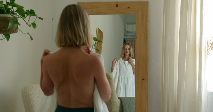 a blonde girl in shorts with a bare back looked into a large mirror and tries on a beautiful white dress in the room. medium shot