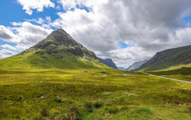 The A82 road crosses pasture fields in the valley floor of Glen Coe. The A82 road crosses pasture fields in the valley floor of Glen Coe, under the mountains of the West Highlands of Scotland. - obrazy, fototapety, plakaty