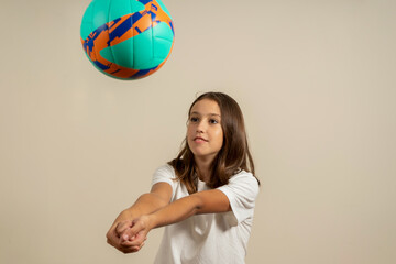 Pretty teen girl with brunette hair making a catch of a volleyball ball with outstretched arms, isolated on beige background.