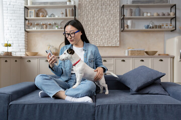 Young beautiful asian woman sitting on sofa at home and using phone happily, teenager with pet jack...