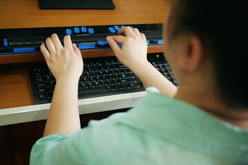 Rear view of person with blindness disability using computer keyboard and braille display or...