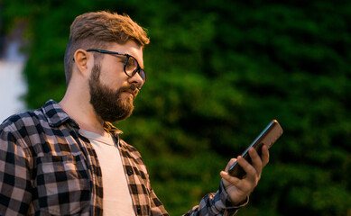 Bearded man wearing eyeglasses is holding scrolling texting in his cellphone at night street. Guy calls for taxi in an app in evening city - copy space and place for advertising