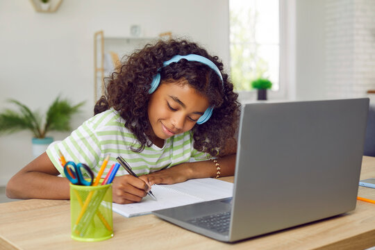 Happy elementary school child studying online. African American student girl in headphones sitting at desk with laptop computer at home, writing in notebook, having remote class or listening to music