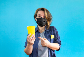 A forty-year-old woman in a shirt points to a yellow cup of coffee. Portrait of a woman in a black medical mask. Woman over isolated blue background