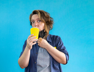 A woman takes off a black medical mask and drinks coffee from a yellow glass on a blue isolated background. Portrait of a woman in a shirt