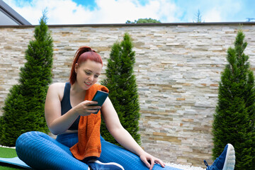 Attractive young fitness girl looking at her smart phone sitting on a pilates ball outdoors. girl exercising while sharing it on her social networks. health and wellness concept.