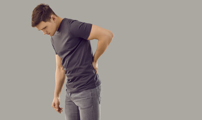 Young caucasian man holding his back because of back pain standing on gray background. Man with painful facial expression holds his hand on injured part of back due to pain in spine or muscles.