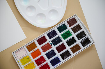 Close-up of colorful palette with refills of watercolour paints and blank white paper sheet with copy space for advertising and promotional text. Painting and drawing activities.