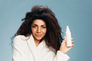 Enjoyed curly Latin lady in bathrobe with toner spray in hands smiling at camera posing isolated on...