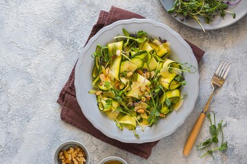 A healthy salad of raw zucchini, cheese, a mixture of microgreens and roasted almond slices with mustard dressing on a gray ceramic plate on a light concrete background. Salad recipes.
