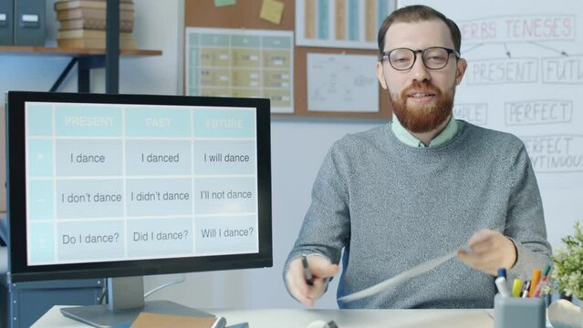 Portrait of young man teaching English online speaking and looking at camera during remote lesson. Teacher pointing at computer screen and showing chart.