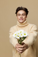 Obraz na płótnie Canvas A handsome young man with fair skin with dark, short, beautiful hair combed back in a beige turtleneck with a high neck stands on a dark beige background with a bouquet of daisies.Vertical studio shot