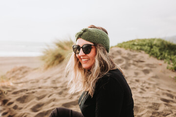 Smiling blonde woman dressed in black, wearing a headband, sunglasses and sitting on the beach. Overcoming depression