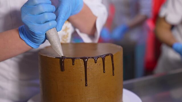 Cake decoration with chocolate. Close up. Gloved hands of confectioner squeeze liquid melted chocolate glazing on the cake edge.
