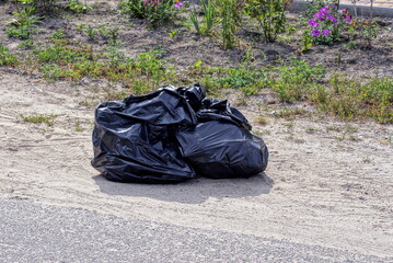 two black plastic bags with garbage stand on the gray ground on the street near the asphalt road