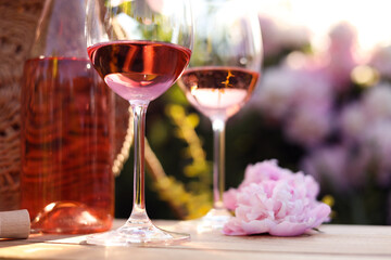 Bottle and glasses of rose wine near beautiful peony flower on wooden table in garden, closeup