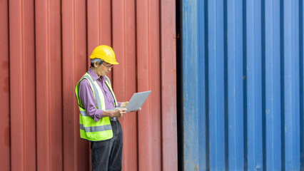 Elderly manager in safety uniform uses computer notebook and technology to check stocks in a logistic cargo. Old man with hard hat and safety jacket is using laptop next to a blue container